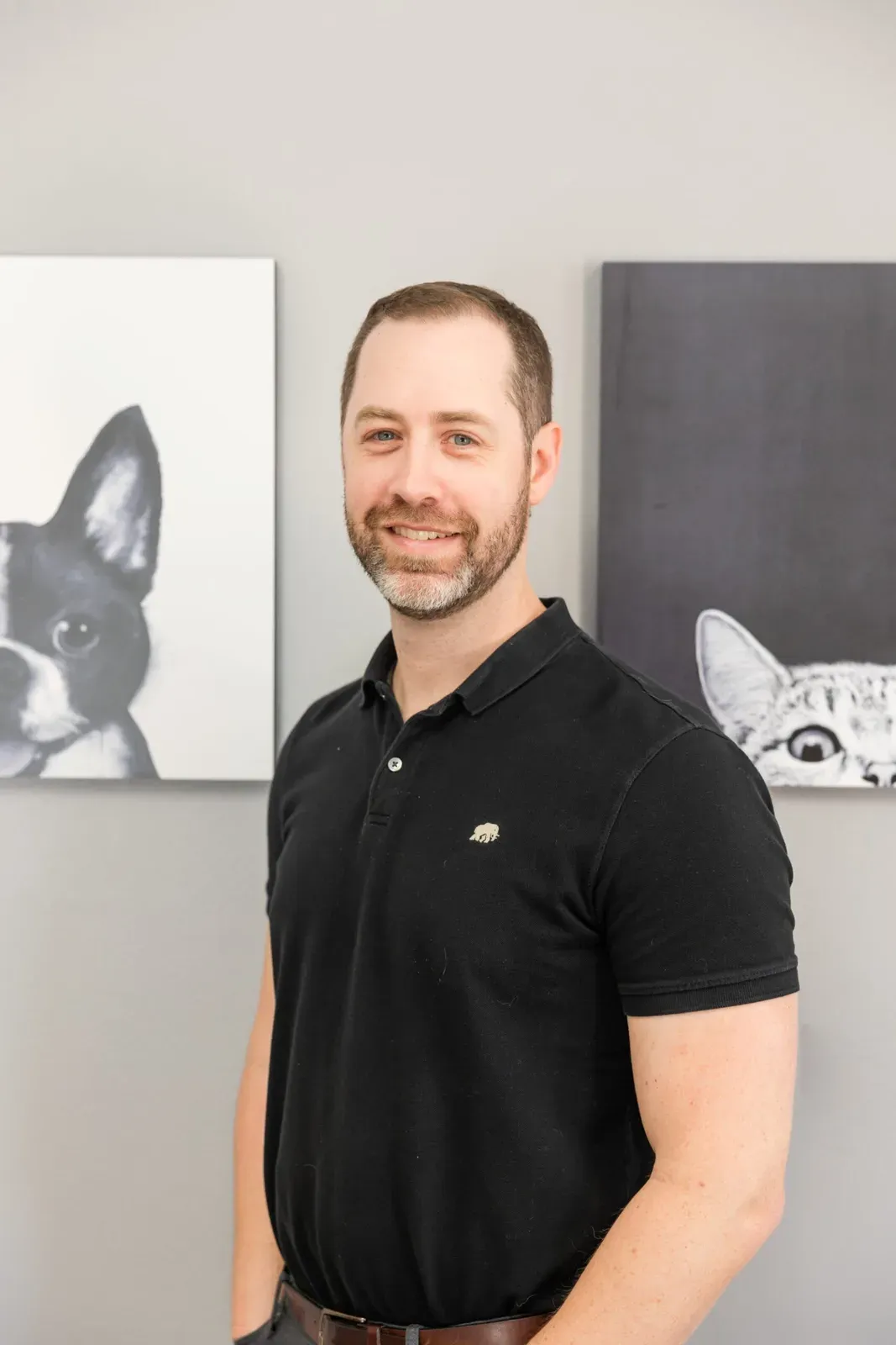 A staff member in a black polo shirt smiles beside framed dog and cat artwork