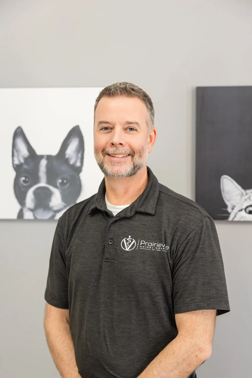 A smiling staff member in a hospital polo shirt stands before dog and cat artwork