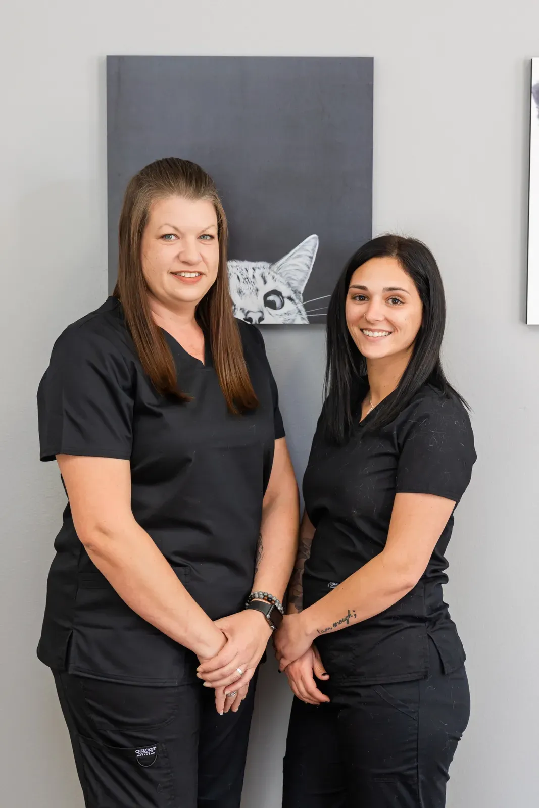 Two staff members in black scrubs smiling in front of framed cat artwork
