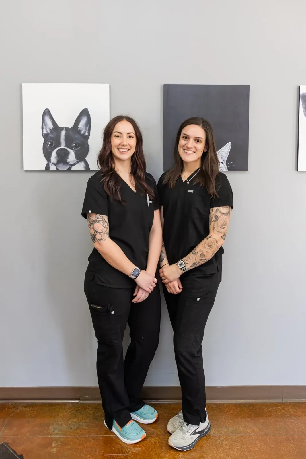 Two staff members in black scrubs stand smiling beside framed pet portraits