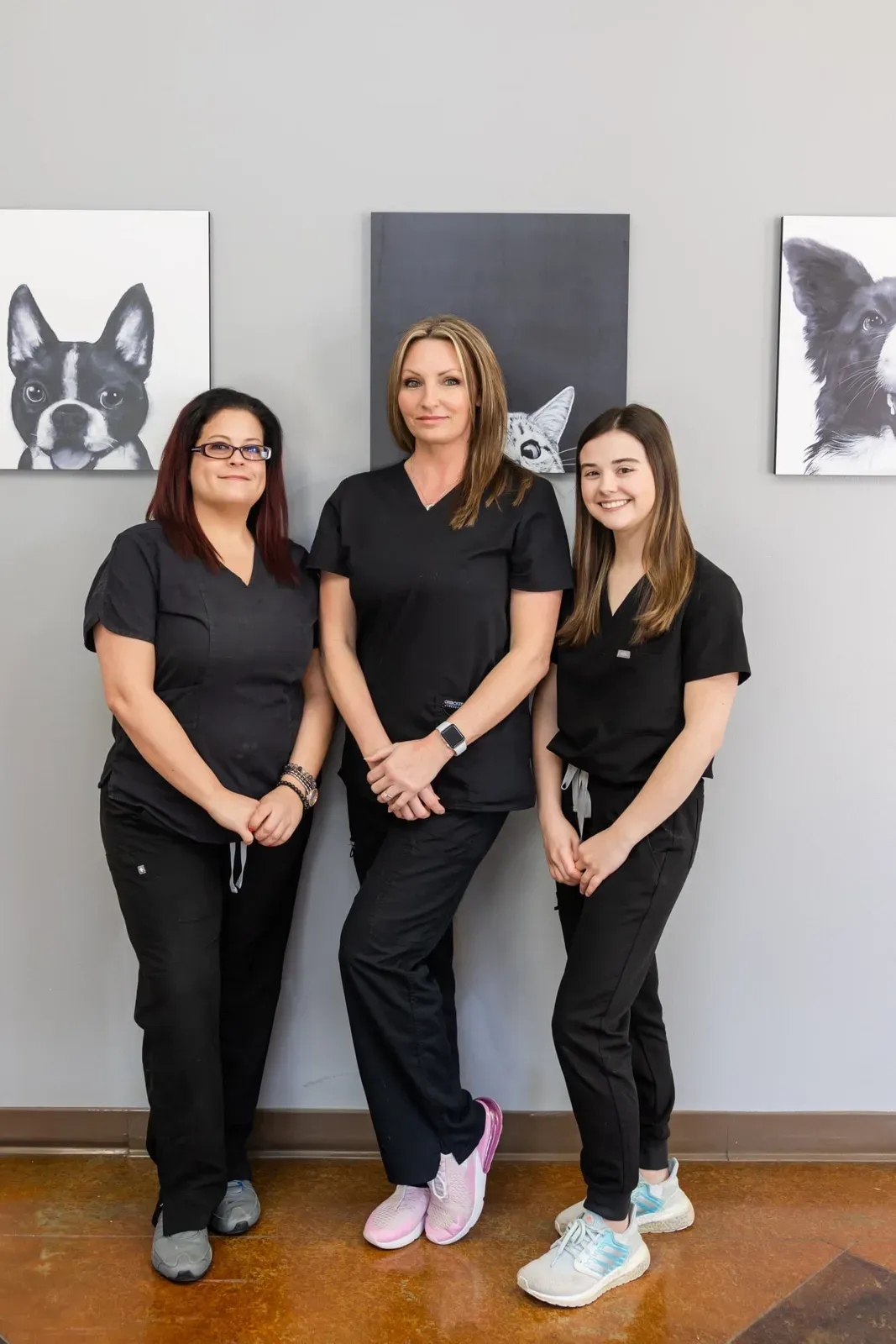 Three staff members in black scrubs stand together in front of pet artwork